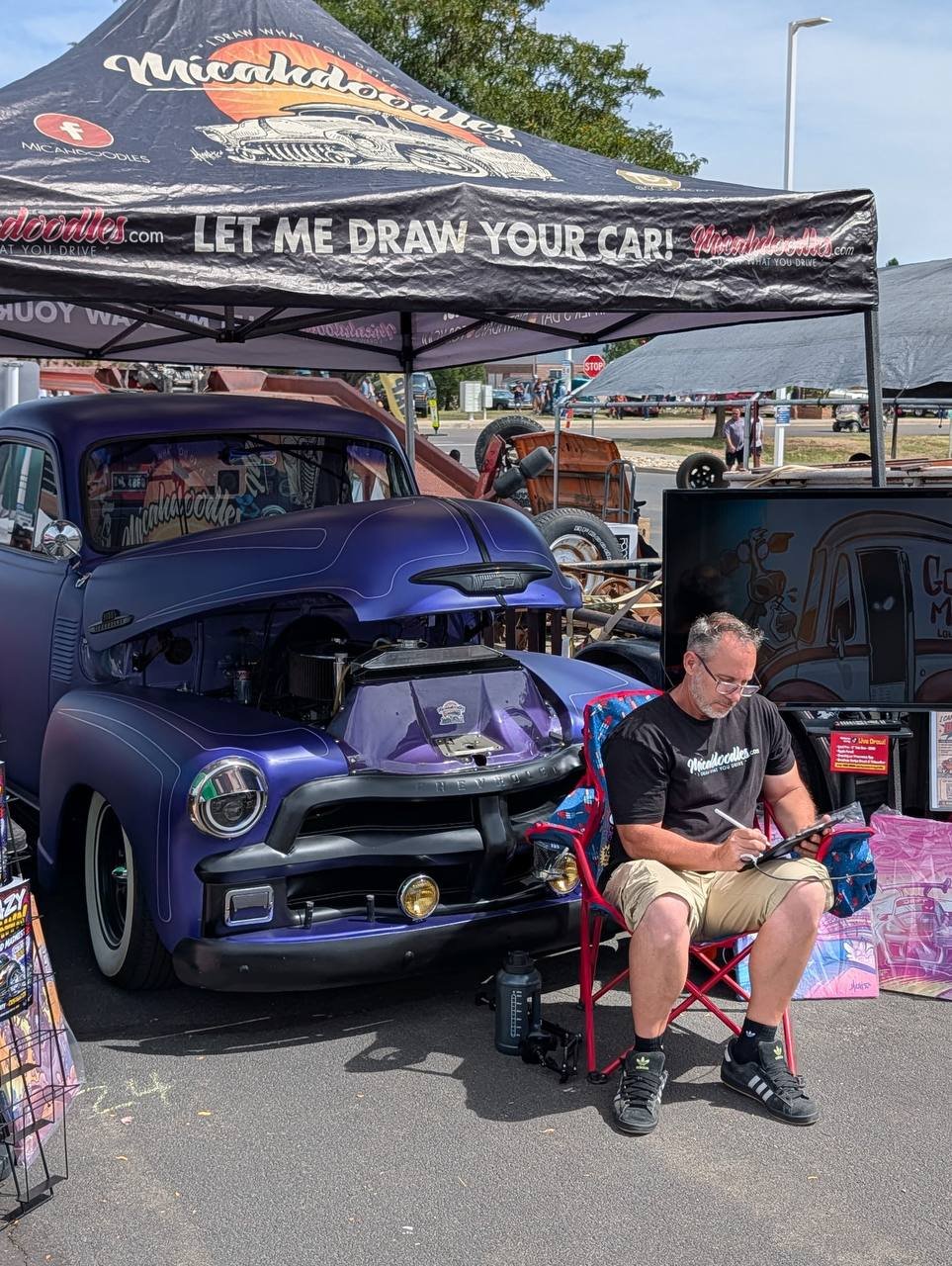 Micah drawing a custom CARtoon at a car show beside a purple hot rod truck